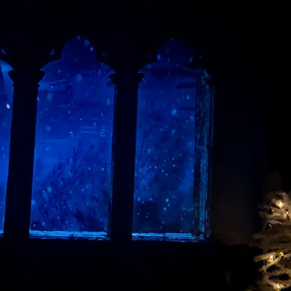 Three arched castle windows with blue snowy scenes and a lit Christmas tree beside them.